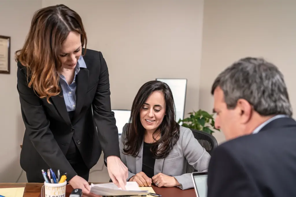 Sara standing next to Michelle pointing at paperwork and Sanford sitting across from them.